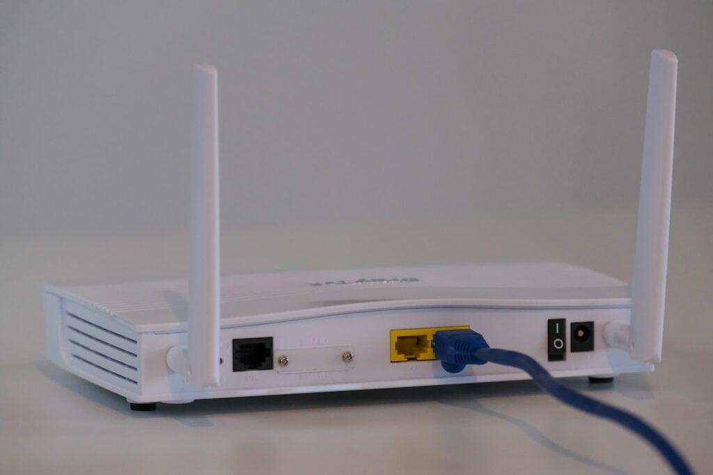 A person plugs an Ethernet cable into a laptop while a Wi-Fi router sits nearby on a desk.