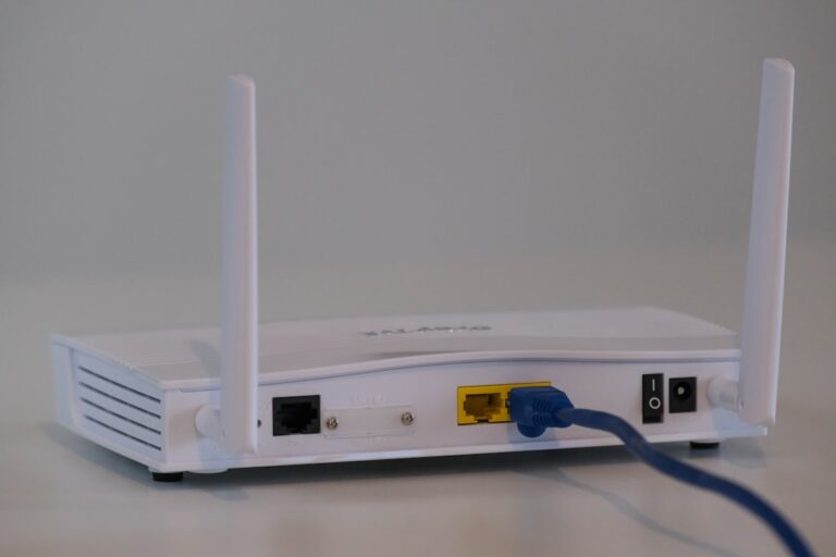 A person plugs an Ethernet cable into a laptop while a Wi-Fi router sits nearby on a desk.